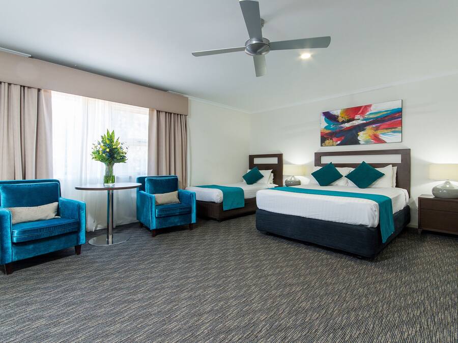 Ein Hotelzimmer mit einem großen Bett, zwei Sesseln, einem kleinen Tisch mit Blumen und einem Deckenventilator. An der Wand hängt ein Gemälde.