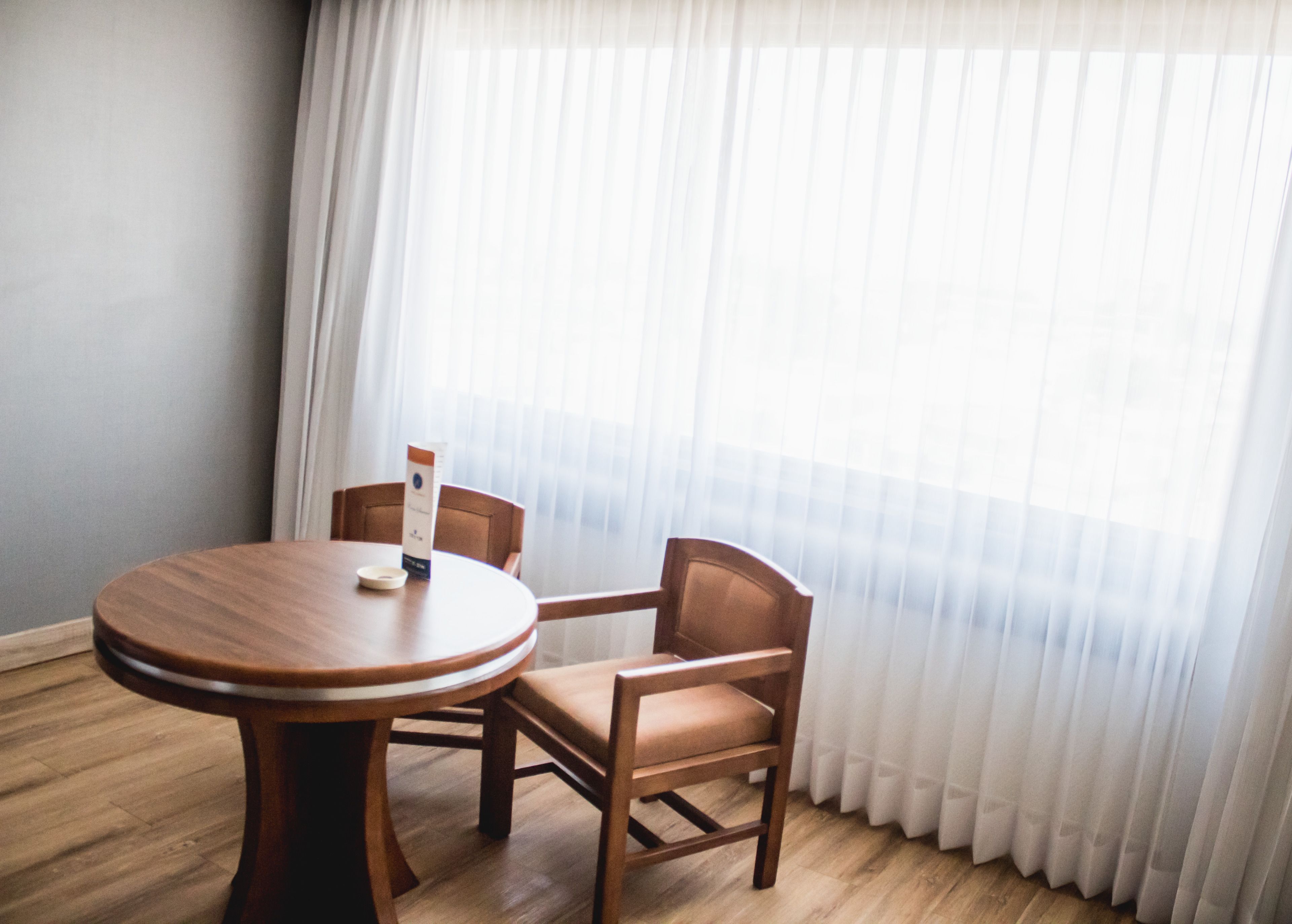 Una habitación con una mesa redonda de madera y dos sillas de madera, una cortina blanca y suelo de madera.