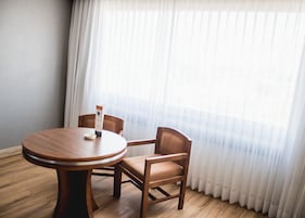 Una habitación con una mesa redonda de madera y dos sillas de madera, una cortina blanca y suelo de madera.