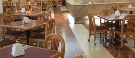 Ein Restaurant mit Holztischen und -stühlen, einer zentralen Theke und einem Fliesenboden.