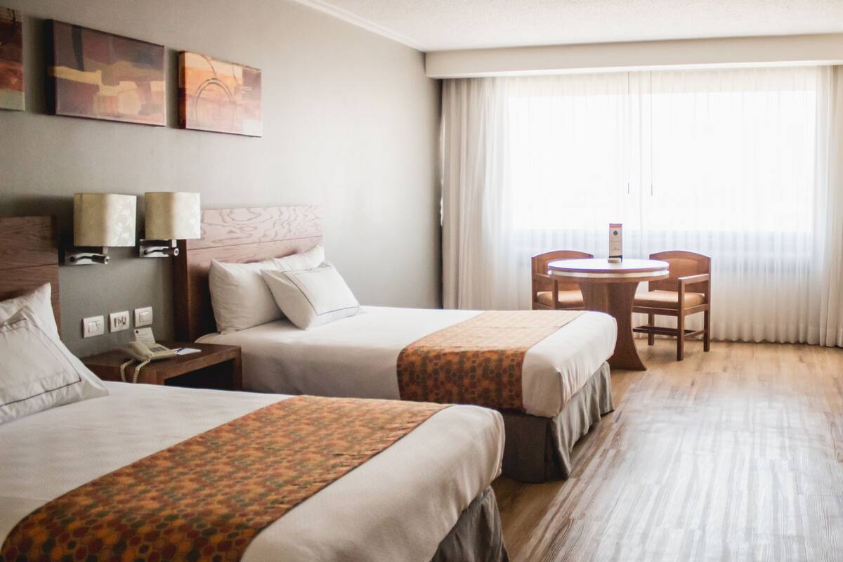 Habitación de hotel con dos camas, una mesita pequeña y una silla. Hay dos lámparas en las mesitas de noche y una obra de arte en la pared.