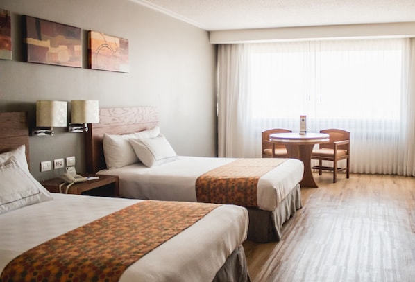 Habitación de hotel con dos camas, una mesita pequeña y una silla. Hay dos lámparas en las mesitas de noche y una obra de arte en la pared.