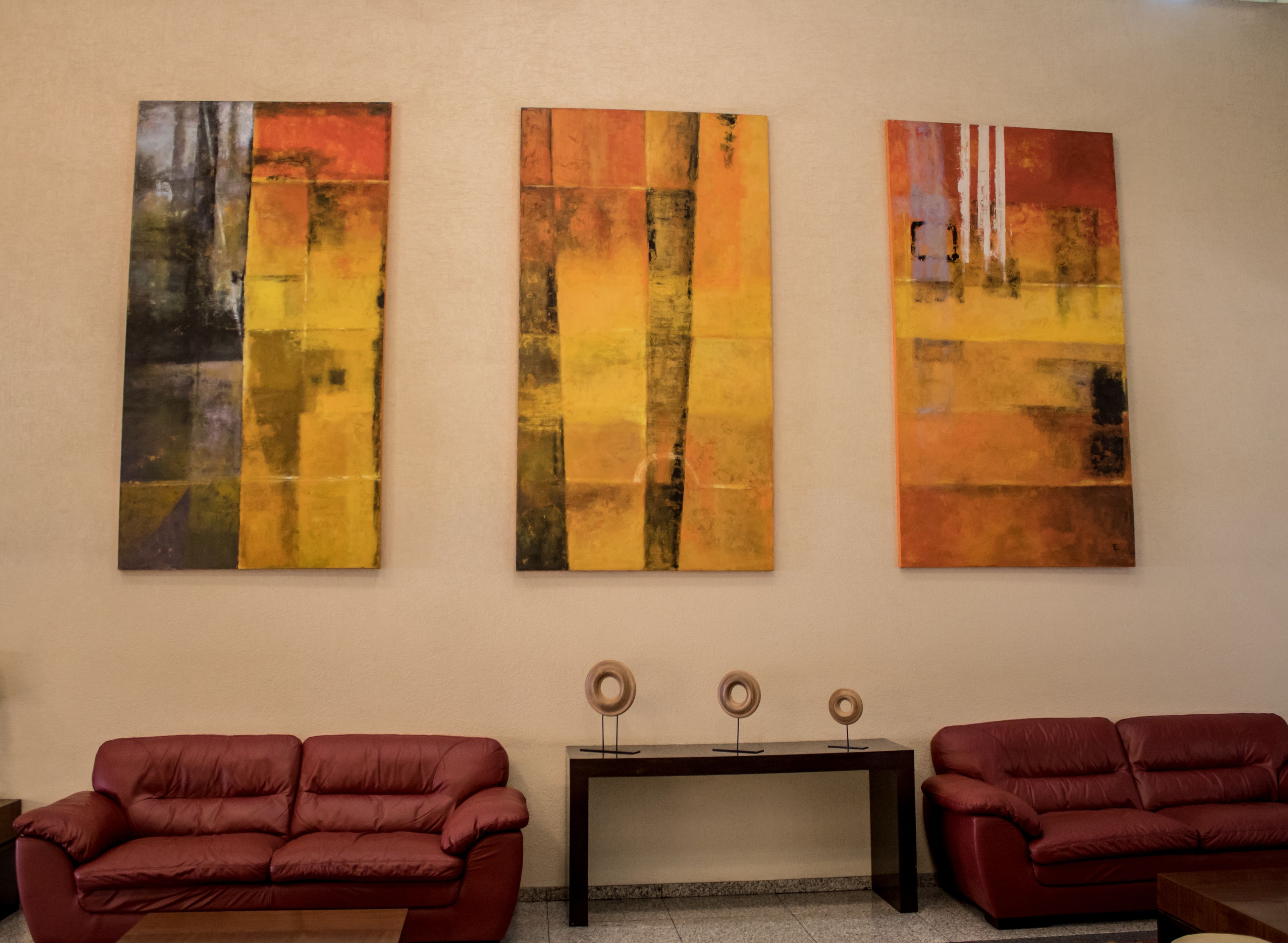 Trois tableaux abstraits accrochés à un mur, une table en bois foncé ornée de trois objets décoratifs et deux canapés en cuir rouge.