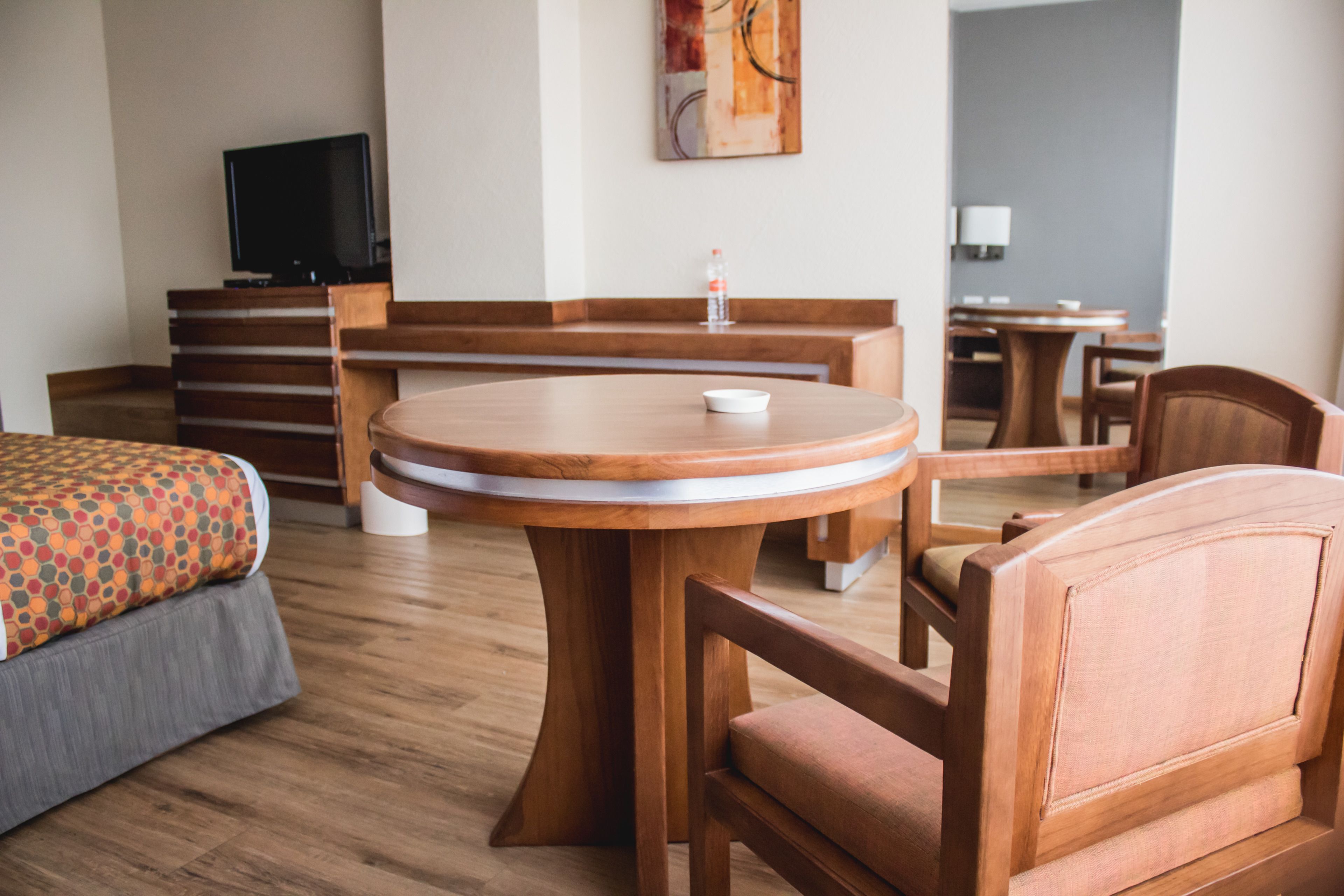 Una habitación de hotel con una mesa redonda de madera, dos sillas de madera, una cama con colcha estampada, un televisor y un cuadro en la pared.
