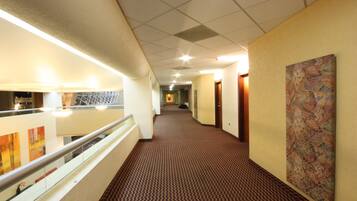 A hotel corridor with a patterned carpet, a glass railing, and a colorful abstract painting on the wall.