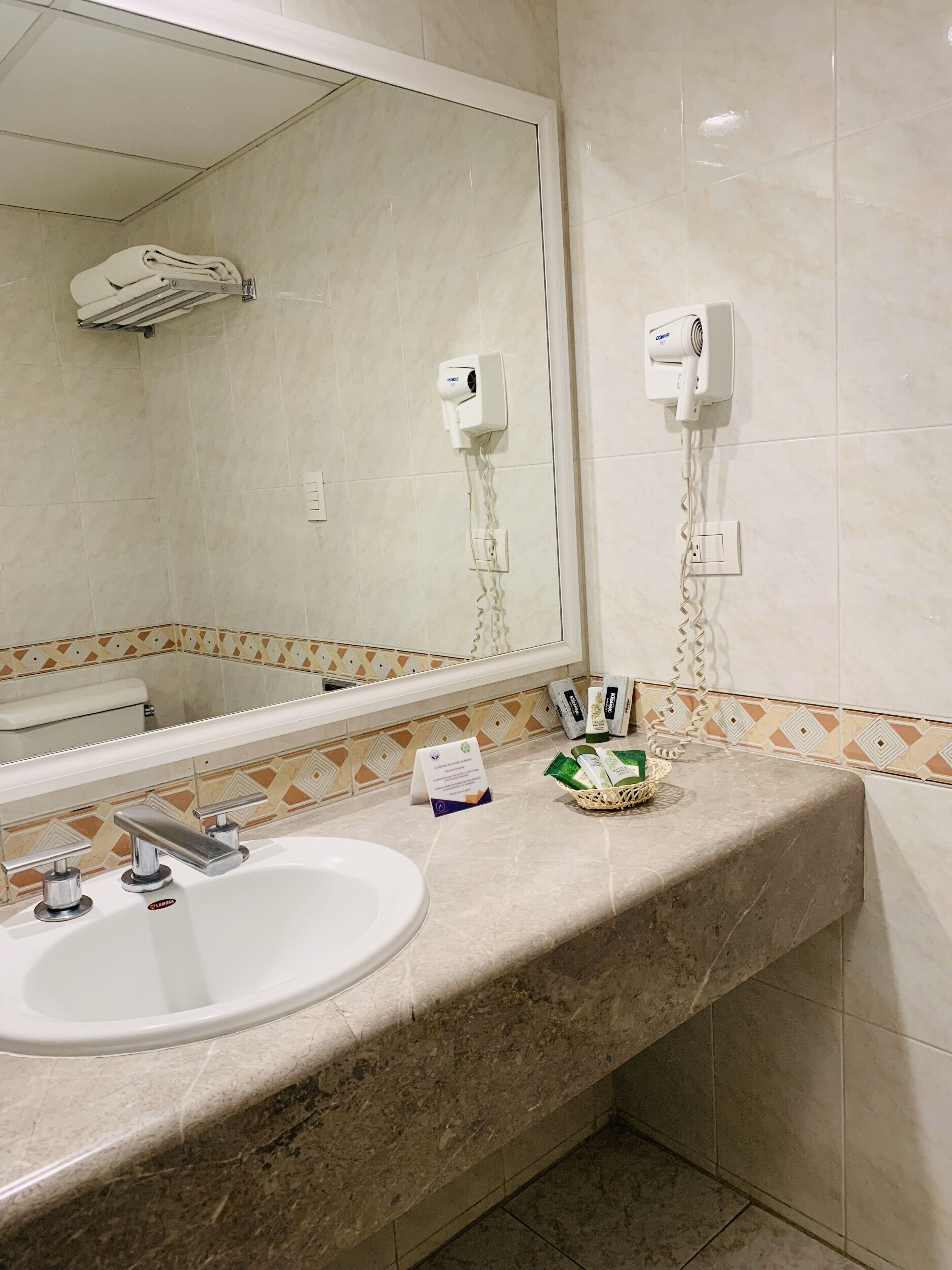 Un baño con un lavamanos blanco, un espejo, un secador de pelo y artículos de aseo en la encimera.