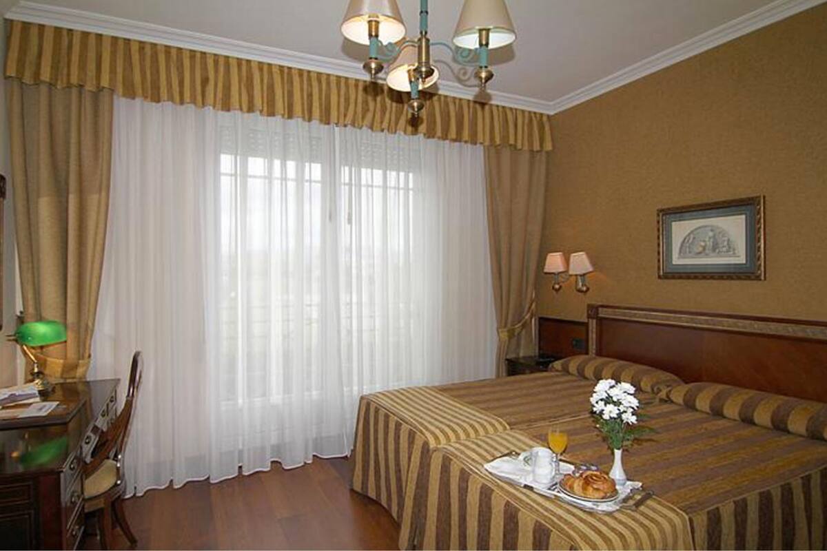 A hotel room with a bed, a desk with a lamp, a chair, a bedside table with a vase of flowers, and a framed picture on the wall.