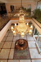 Un gran candelabro con varios niveles y luces, una alfombra estampada, y una pequeña mesa con un arreglo floral en un vestíbulo espacioso con piso de baldosas y barandilla de vidrio.