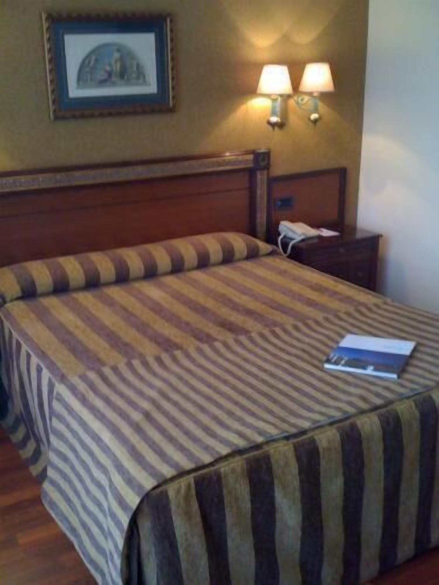 Ein Hotelzimmer mit einem hölzernen Kopfteil, einer gestreiften Bettdecke, zwei Nachttischlampen, einem gerahmten Bild an der Wand und einem Telefontisch.