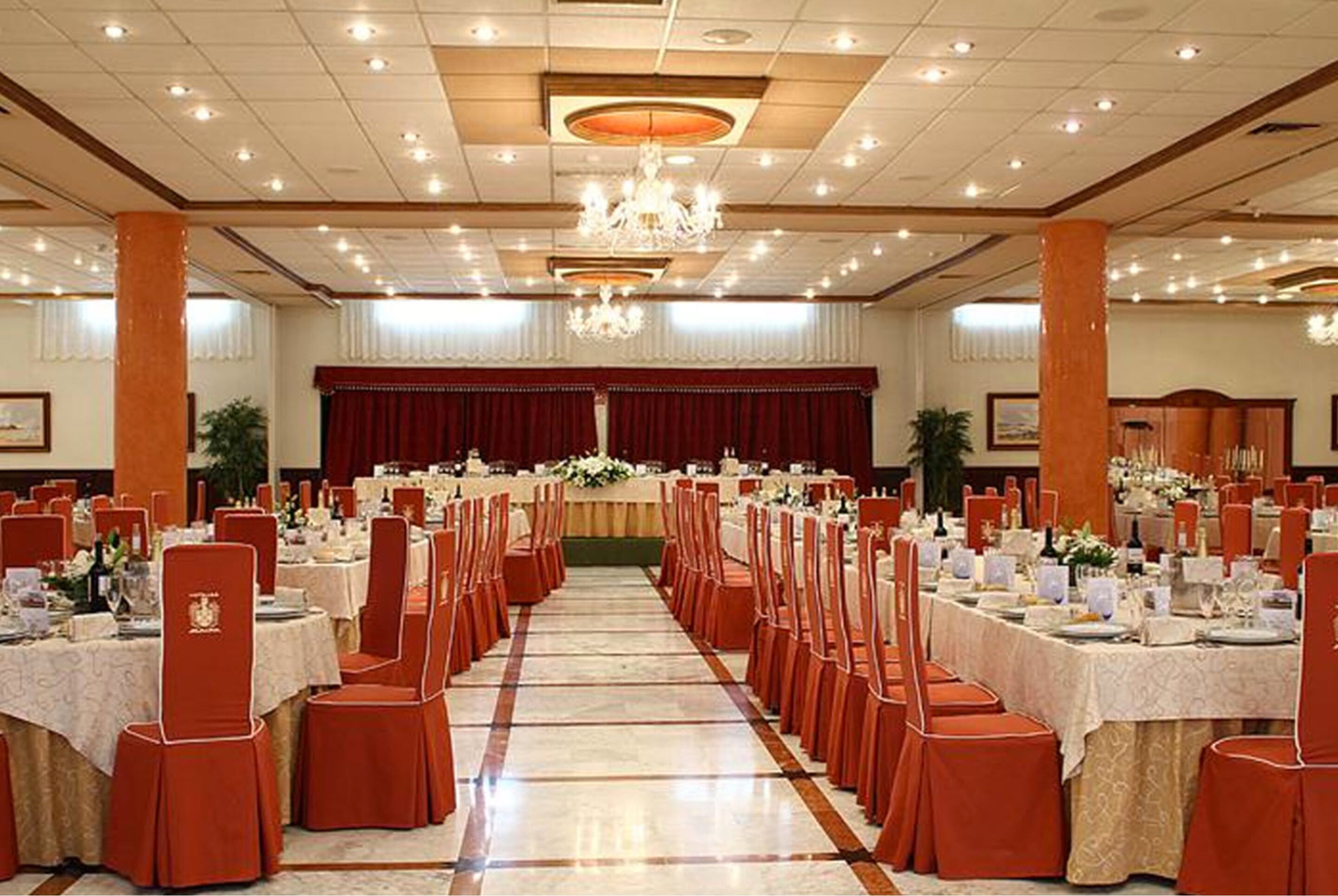 Un salón de banquetes con filas de mesas y sillas, una araña y un escenario con cortinas.