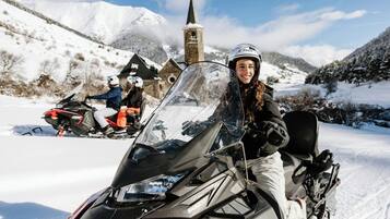 Una moto de nieve con un conductor, una iglesia y montañas nevadas al fondo.