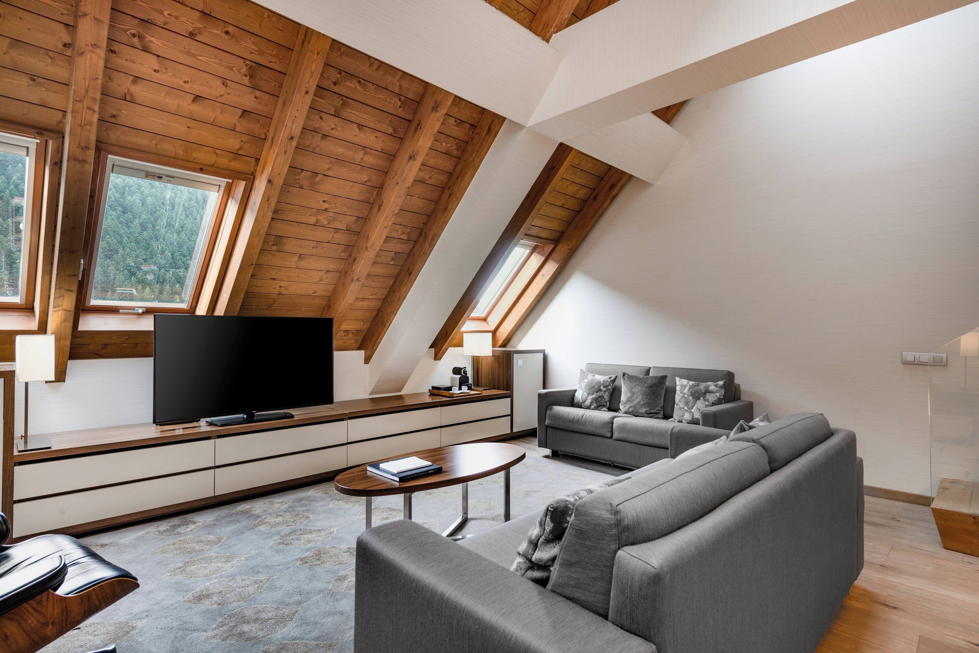 A modern loft apartment with a sloped ceiling, built-in wooden beams, a large flat-screen TV, a grey sofa, and a dining area with a wooden table.