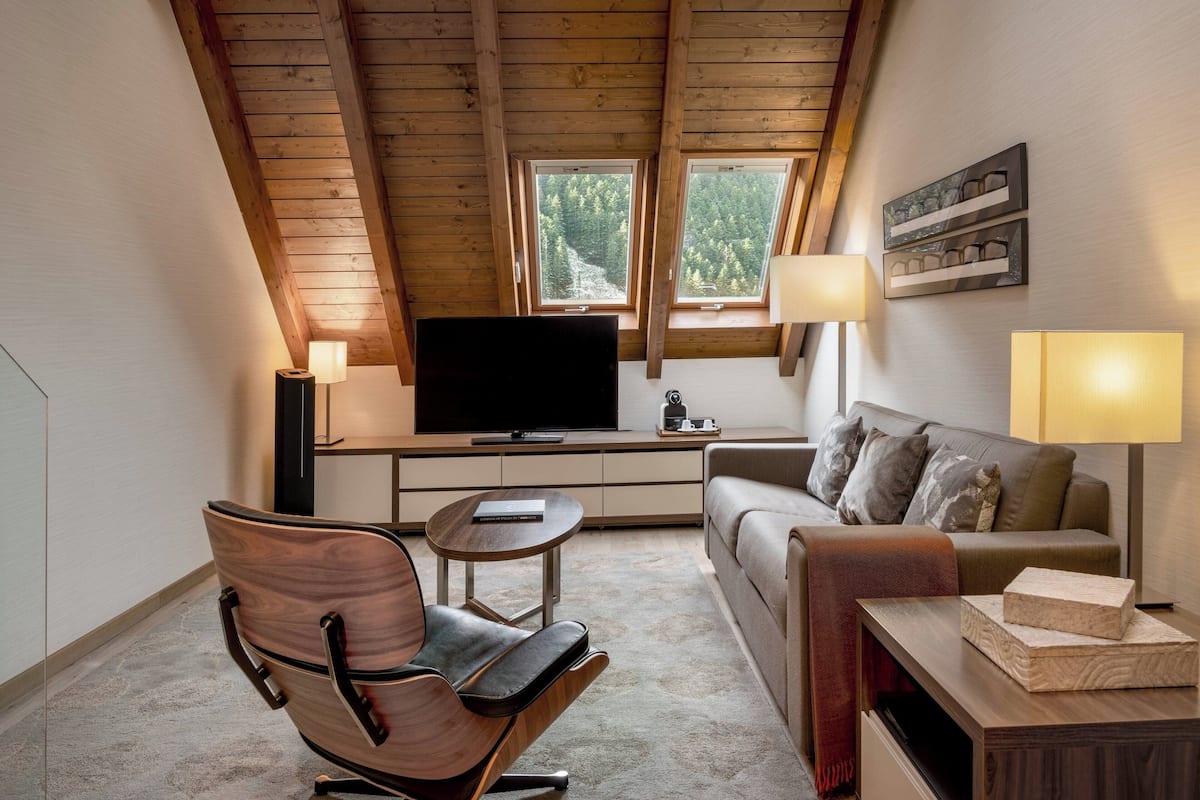 Ein gemütliches Schlafzimmer im Loft-Stil mit Holzbalkendecke, großem Fenster, Flachbildfernseher, Sofa, Sessel und Nachttisch.