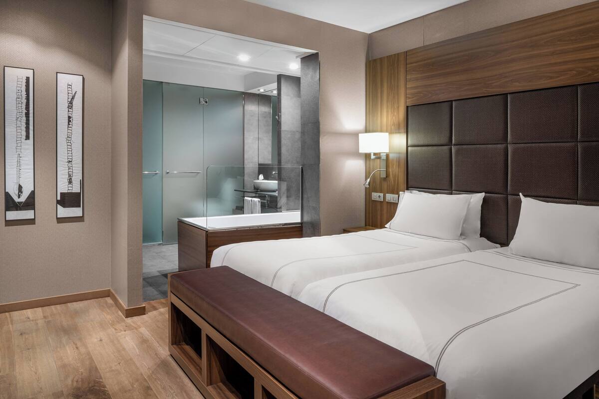 Ein Hotelzimmer mit einem großen Bett, einer Bank, einem Badezimmer mit einer Glasdusche und einem hölzernen Kopfteil.