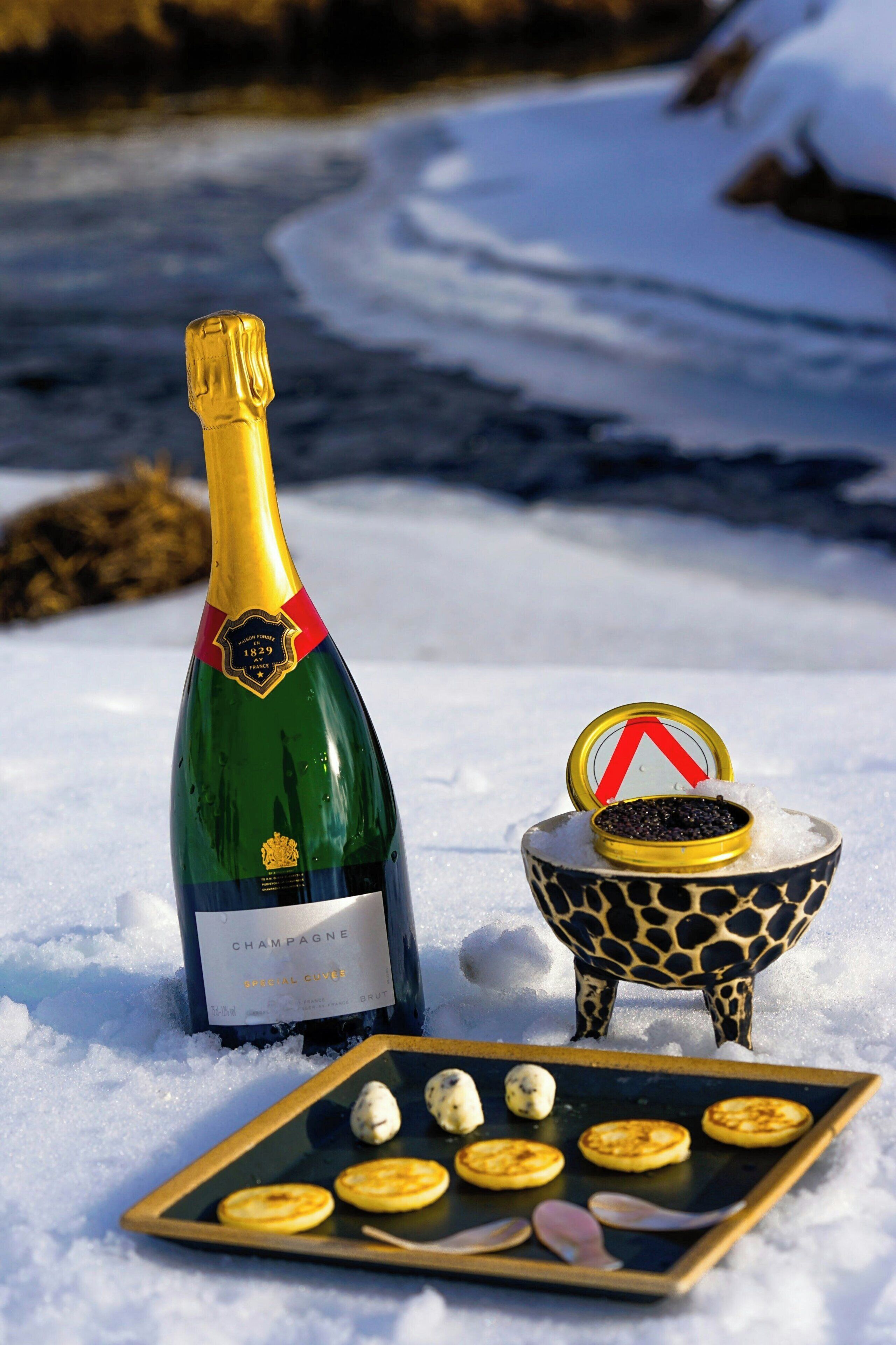 Eine Champagnerflasche, eine kleine Schale mit Kaviar und ein Teller mit verschiedenen Speisen auf einer verschneiten Fläche.