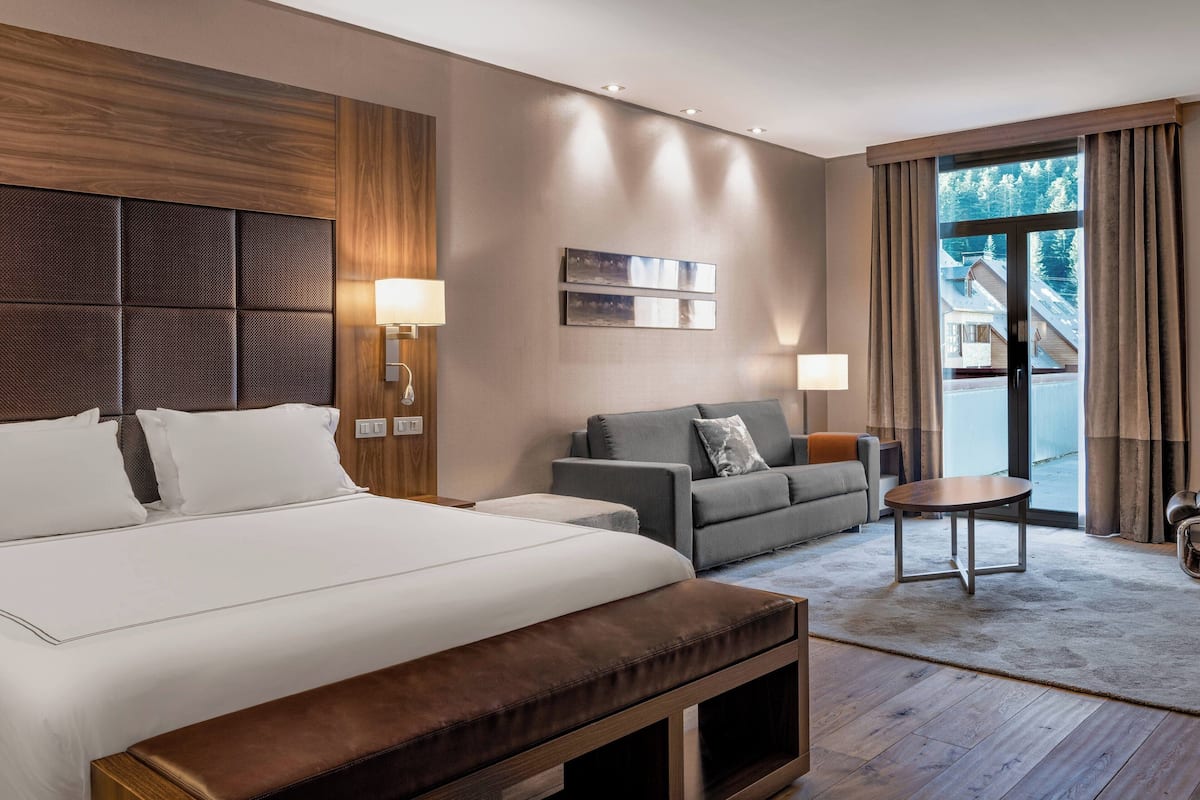 Ein modernes Hotelzimmer mit einem großen Bett, einer Sitzecke, einem kleinen Tisch und Blick auf ein Gebäude durch das Fenster.