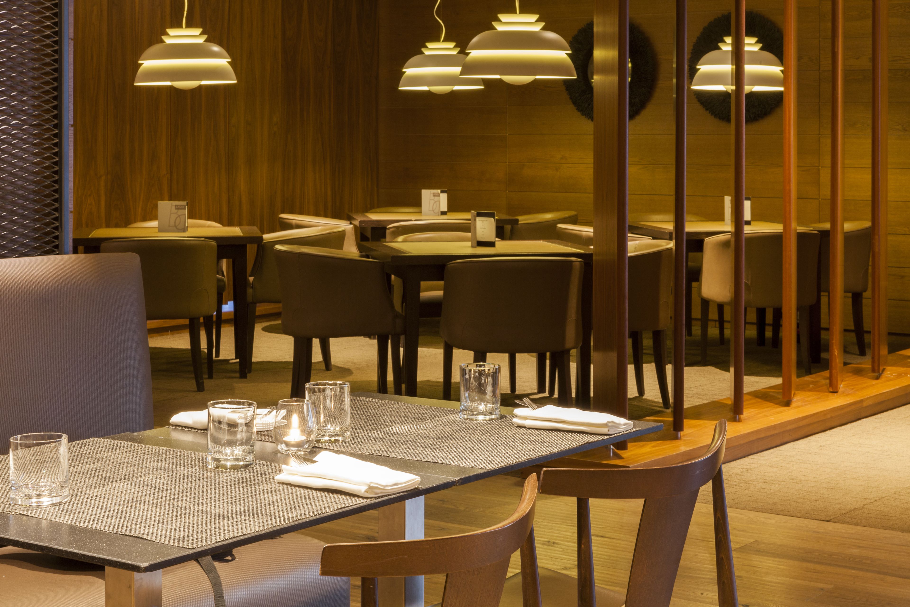 A restaurant interior with multiple tables and chairs, pendant lighting, and a wooden wall.
