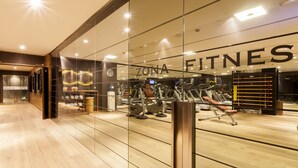 Une salle de sport équipée de matériel d’exercice, dotée d’un plancher en bois et d’un mur en verre sur lequel on peut lire les mots « ZONA FITNESS ».
