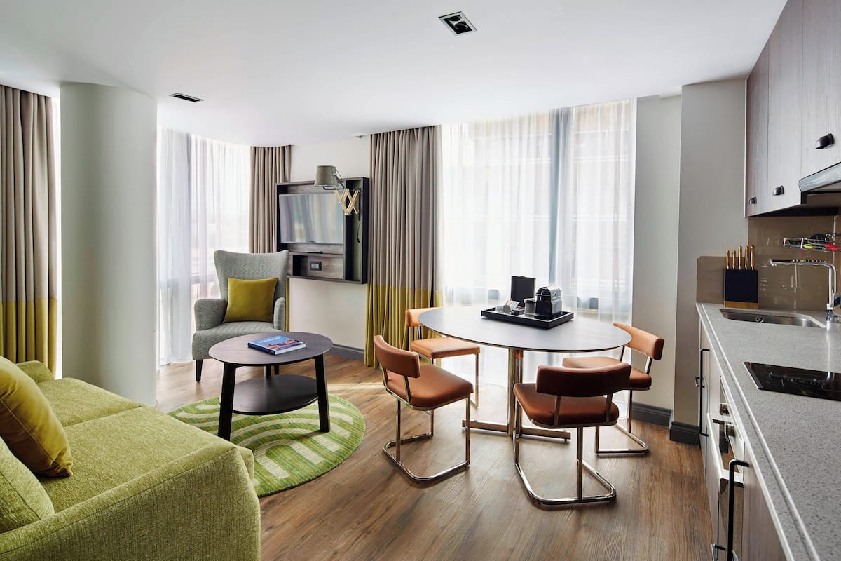A modern hotel room with a green sofa, a round dining table with four chairs, a kitchen area with a sink and stove, and a television on a stand.