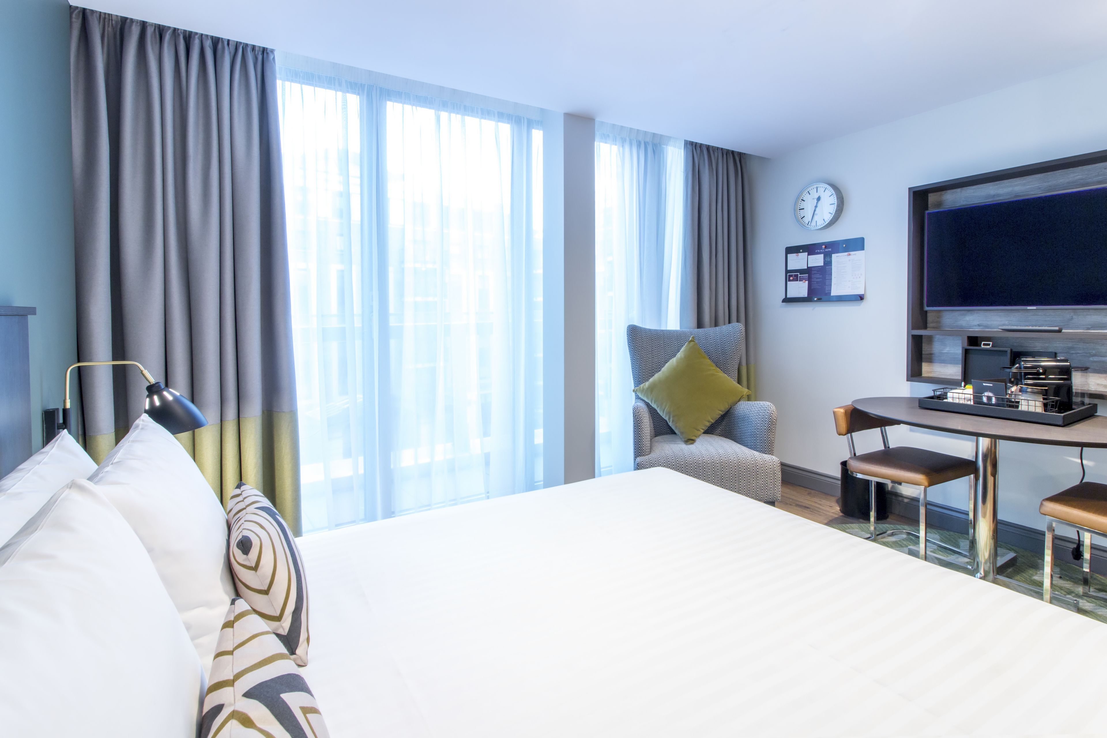A hotel room with a large bed, a desk with a chair, a television, and a clock on the wall.