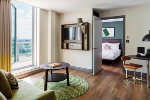 Ein Hotelzimmer mit großem Fenster, einem Bett, einem Schreibtisch mit Stuhl, einem kleinen Tisch und einem an der Wand befestigten Fernseher.
