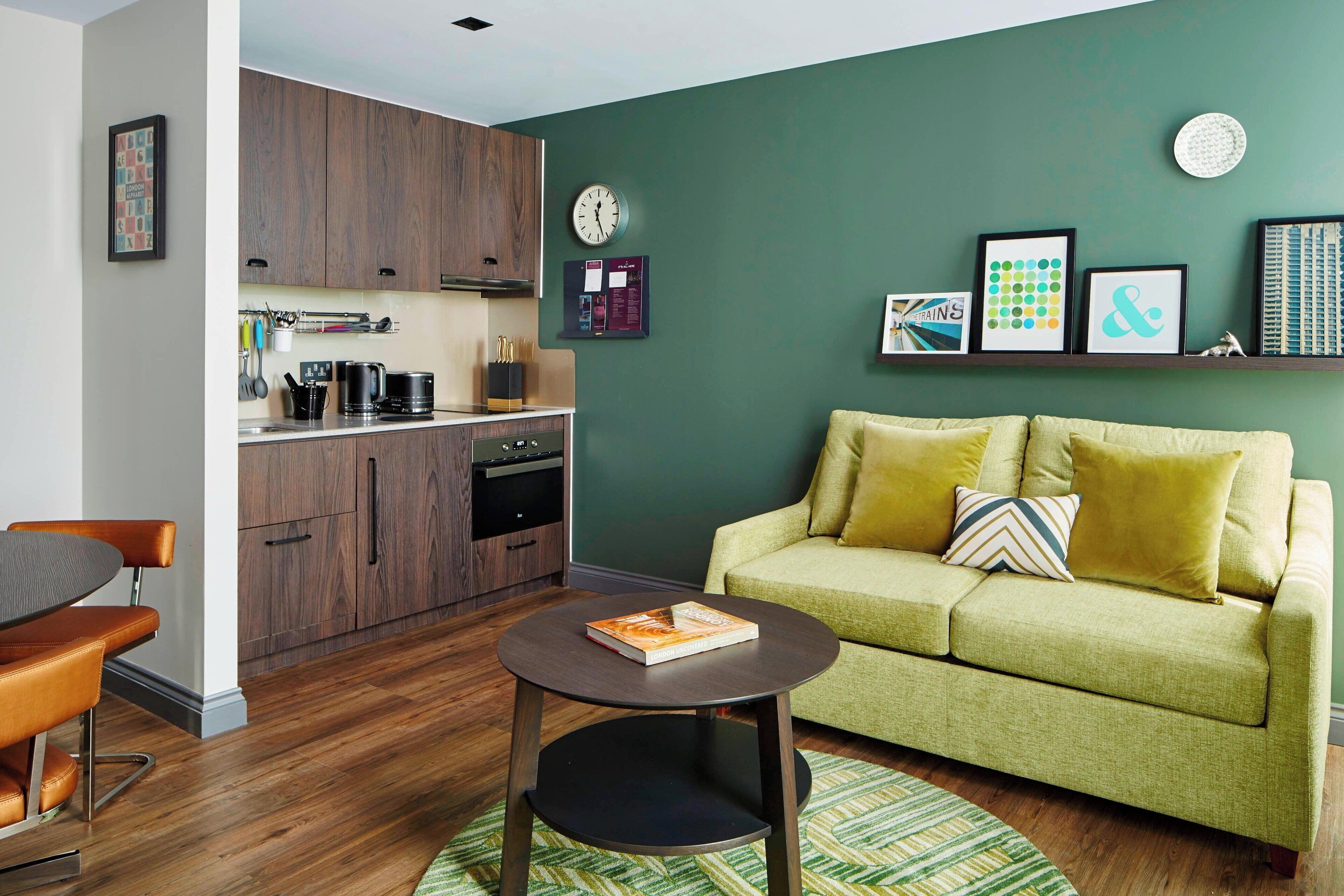 Uma sala de estar moderna com um sofá verde, uma mesa de centro redonda e uma área de cozinha com armários de madeira e um relógio.