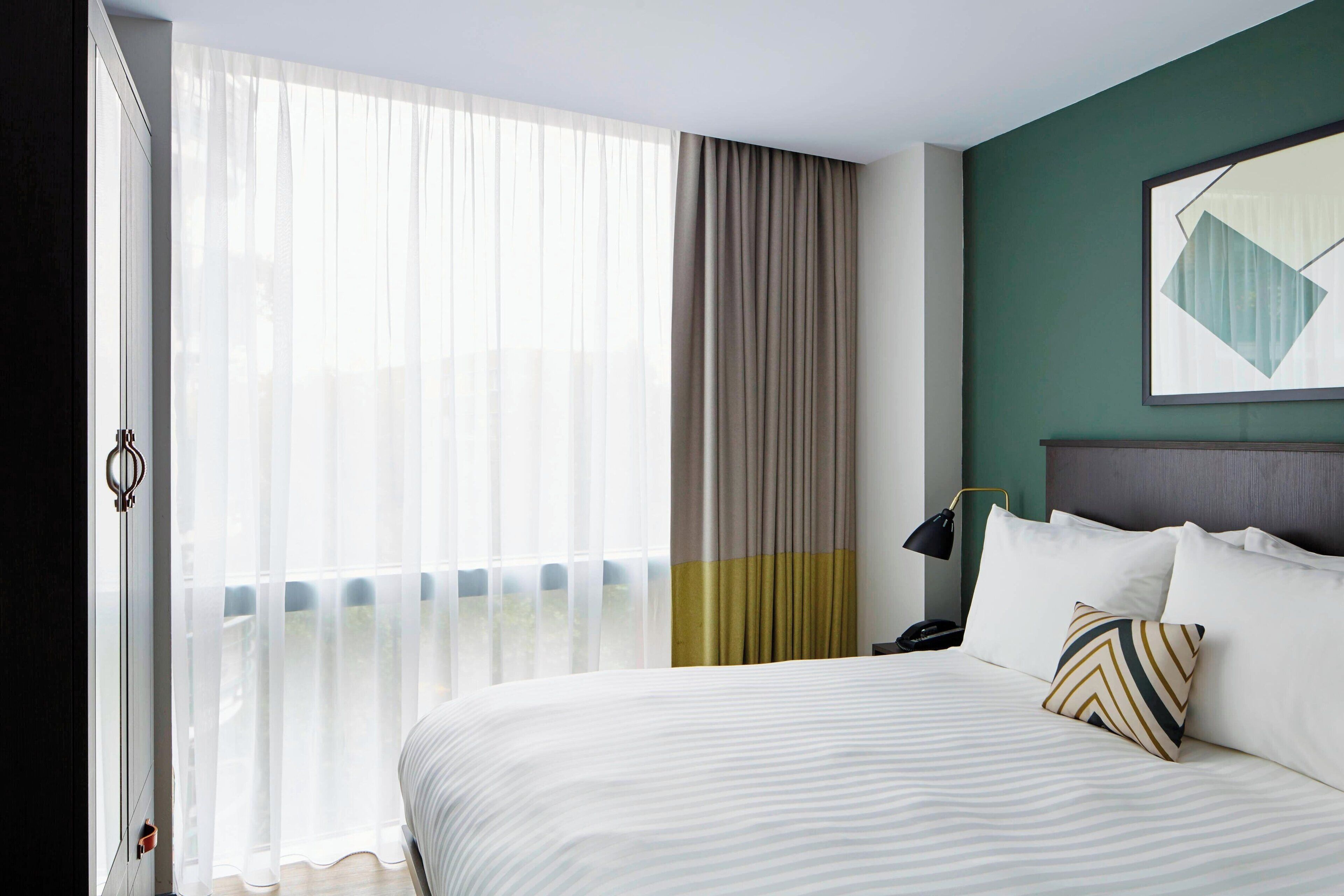 A hotel room with a large bed, a view through sheer curtains, a bedside table with a lamp, and a wall-mounted artwork.