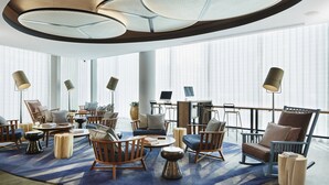 Un área de descanso moderna con diversas opciones de asientos, incluyendo sofás y sillones, dispuestos alrededor de una mesa de centro. Varias lámparas de pie y un ventanal grande ofrecen luz natural.