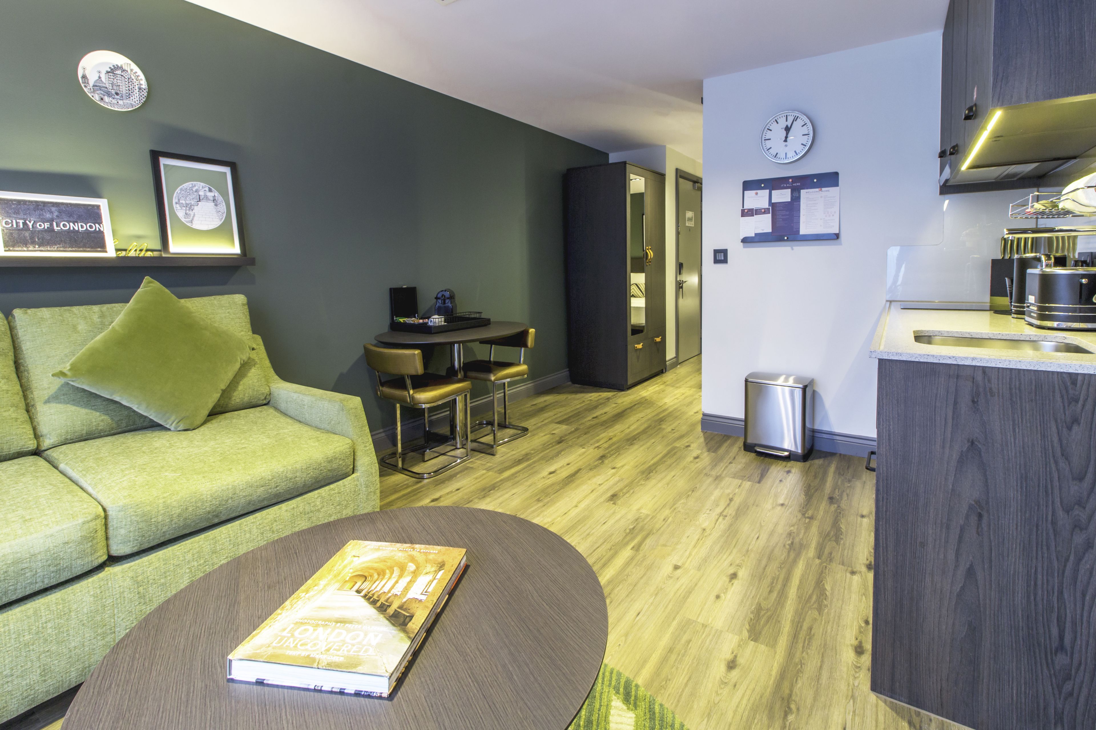 A modern living room with a green sofa, a round coffee table with a book, a dining area with a small table and chairs, and a kitchen with a sink and stove.