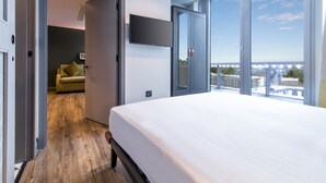 Una habitación de hotel moderna con una cama grande, un televisor montado en la pared y vistas a la ciudad a través de amplios ventanales.