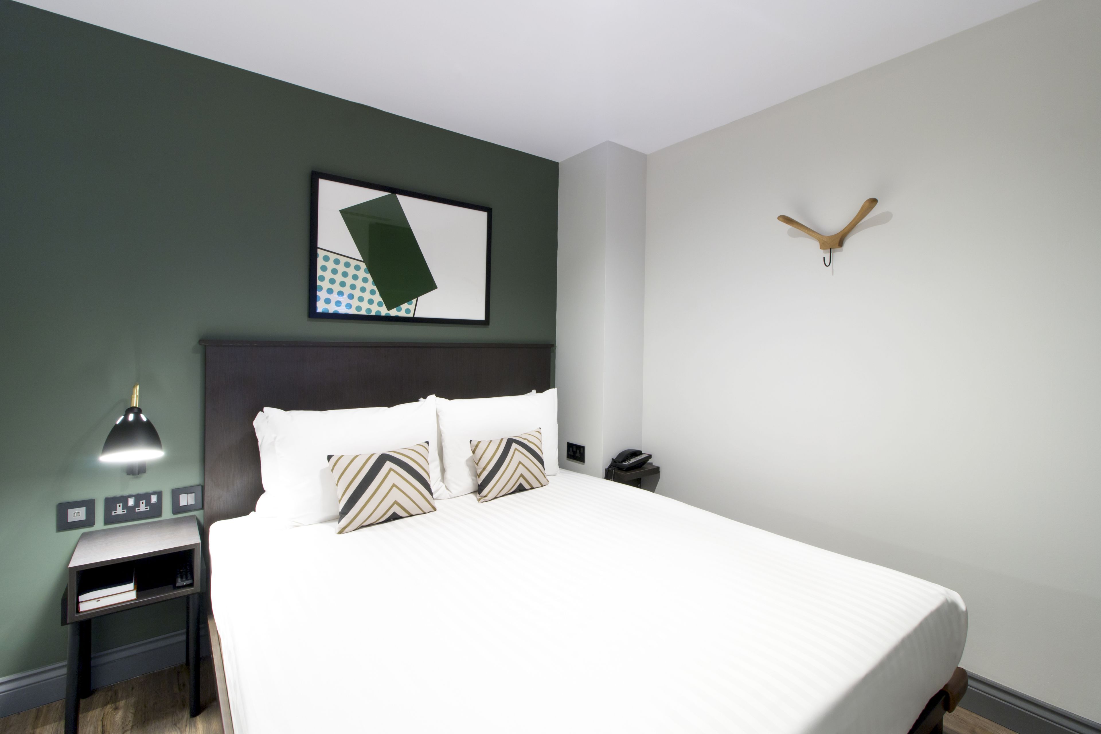 A neatly made bed with a white comforter and striped pillows, a bedside table with a lamp, and a wall-mounted artwork.