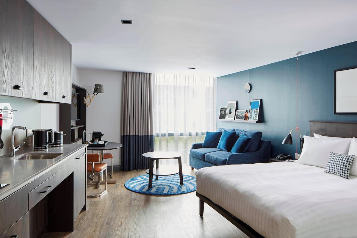 A modern hotel room with a kitchenette, a dining area, a sofa, and a bed.