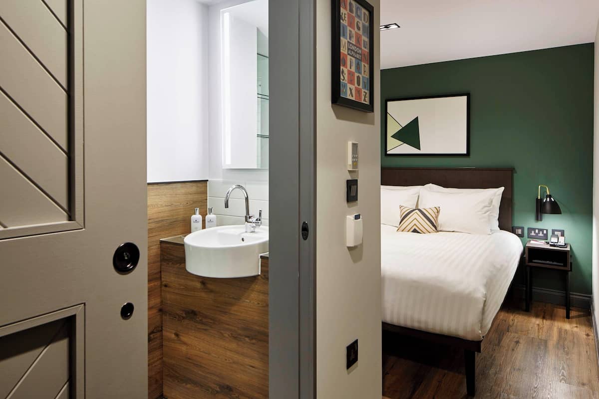 A hotel room with a wooden floor, a white sink, a bed with white bedding, a nightstand with a lamp, and a framed picture on the wall.
