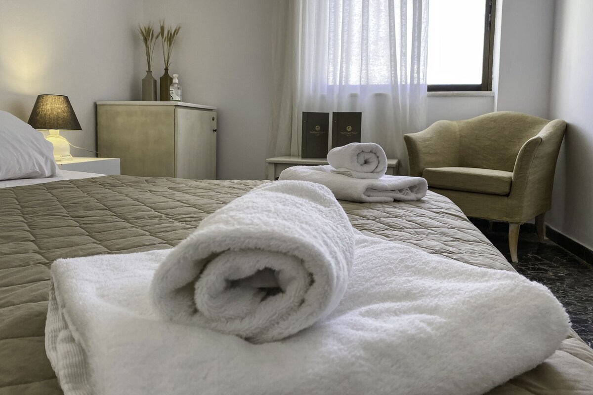 Una habitación de hotel con cama, una silla, una mesita de noche con lámpara y una ventana con cortinas.