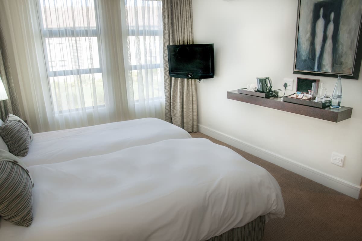 A hotel room with two beds, a flat-screen TV, a mounted shelf with toiletries, and a painting on the wall.