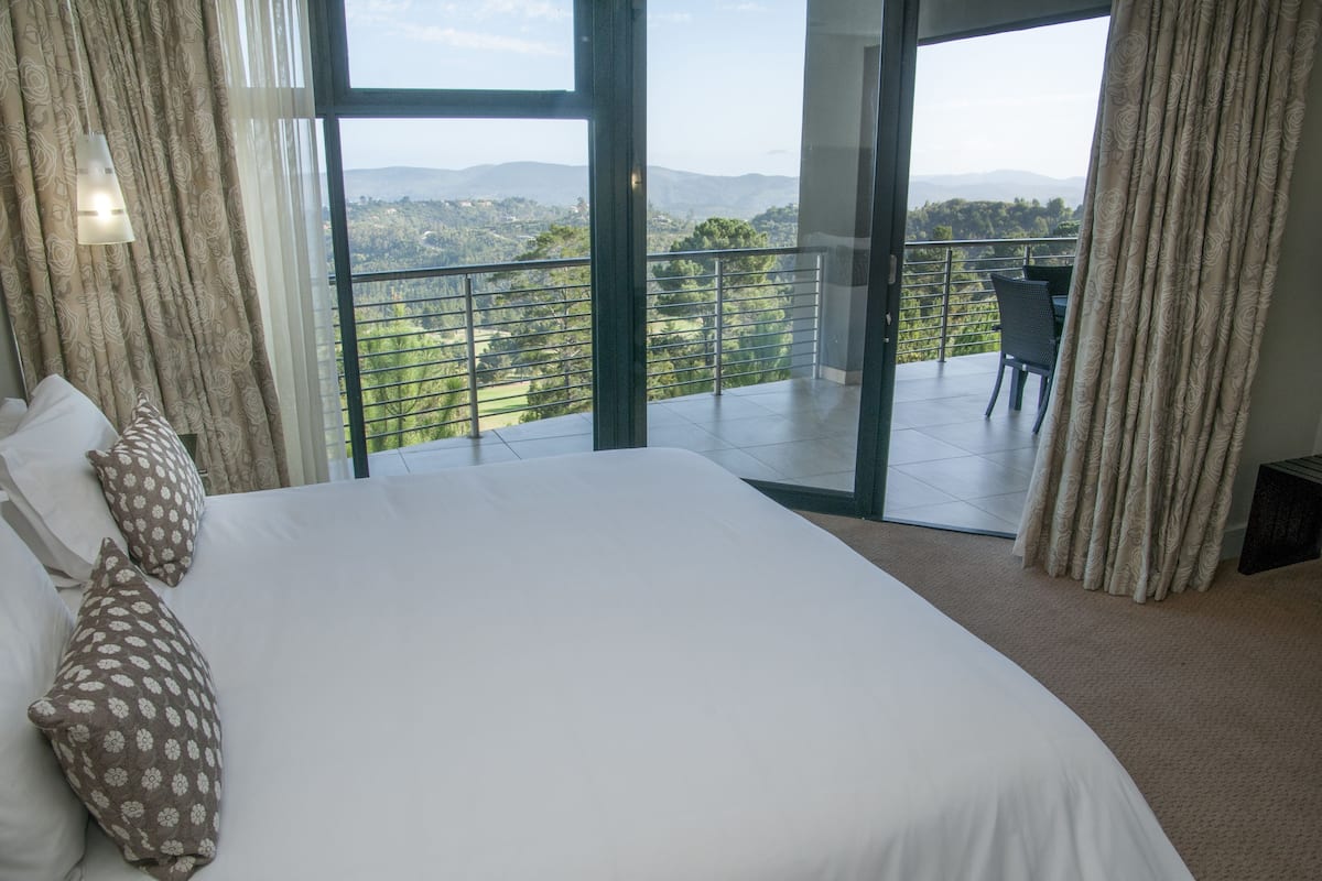 A bedroom with a large bed, a view of a balcony with a table and chair, and a scenic landscape view.