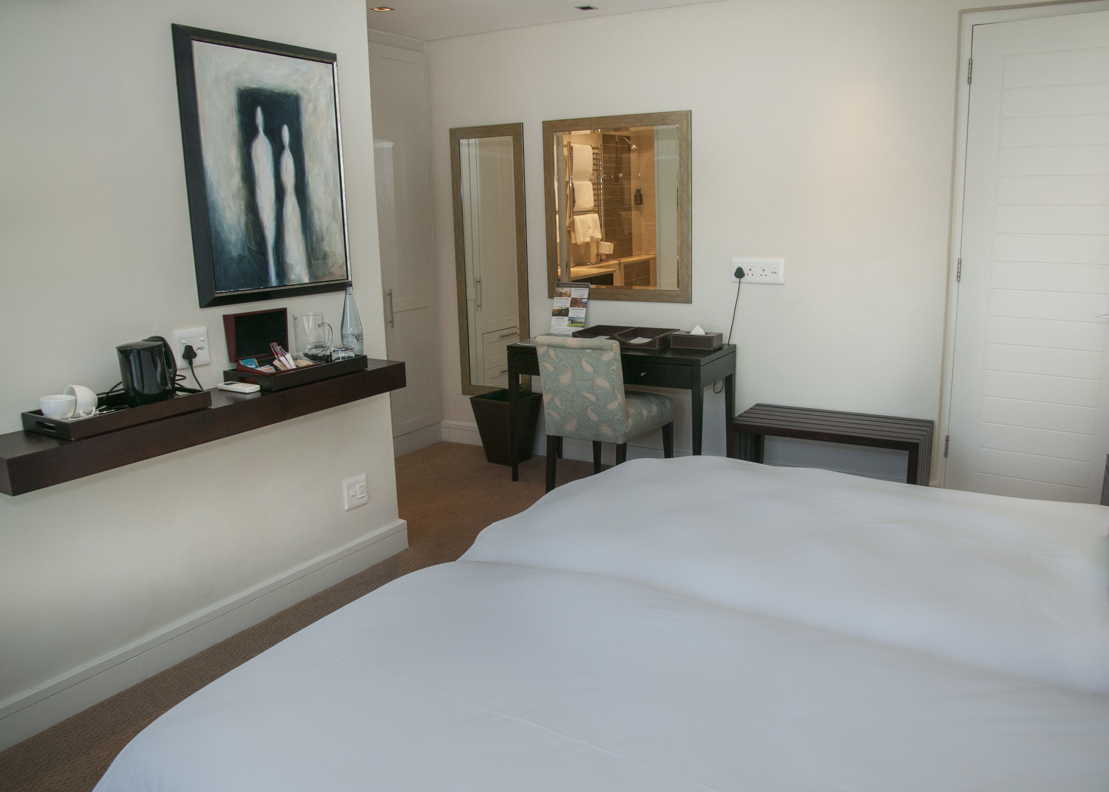 A hotel room with a large bed, a desk with a chair, a coffee maker, a mirror, and a painting on the wall.