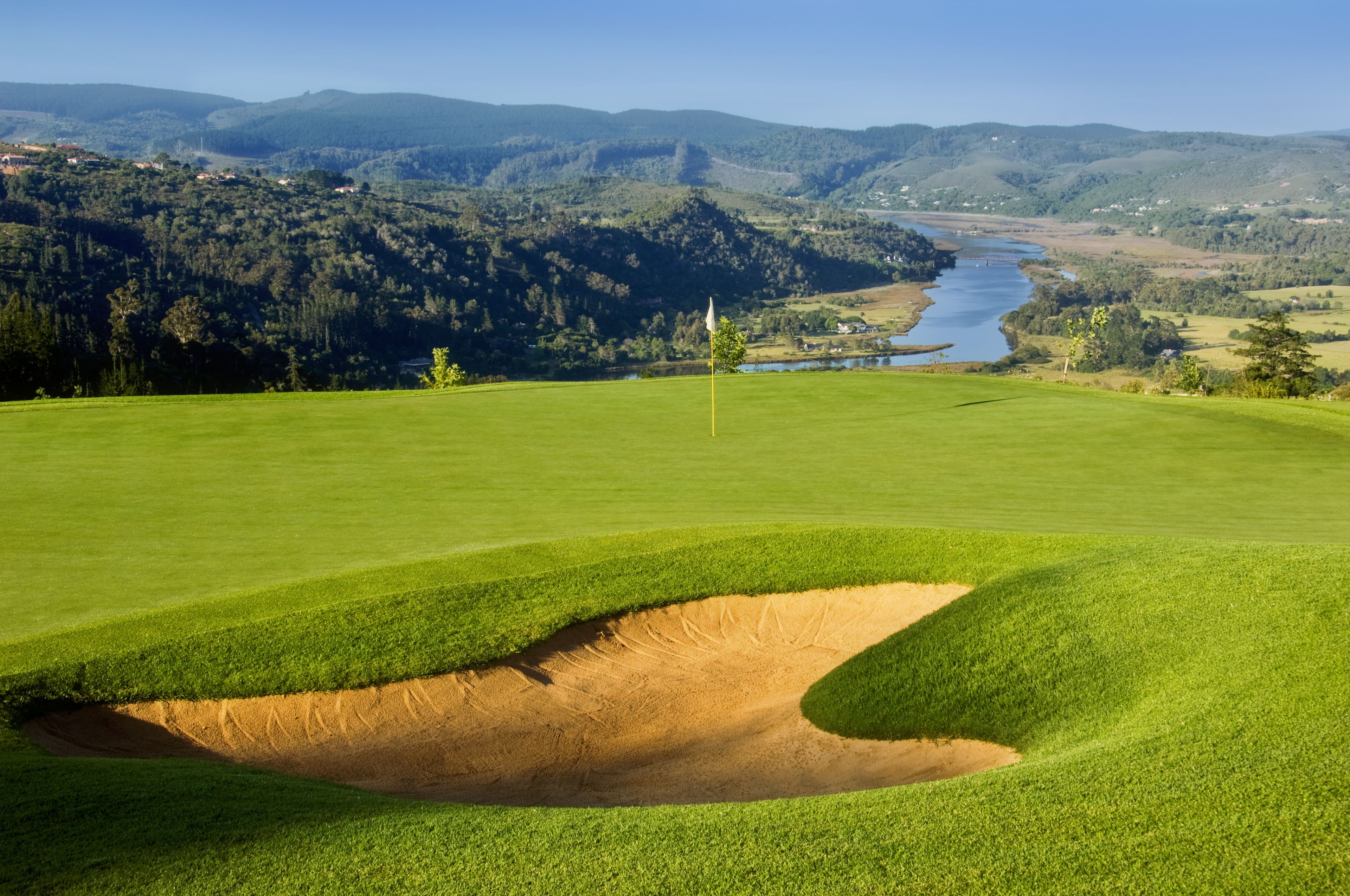 A golf course with a sand bunker, a flagstick, and a river in the background.