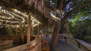 An outdoor wooden deck with benches and tables, surrounded by trees and a thatched roof structure with string lights. - Simola Hotel, Country Club & Spa (Knysna)