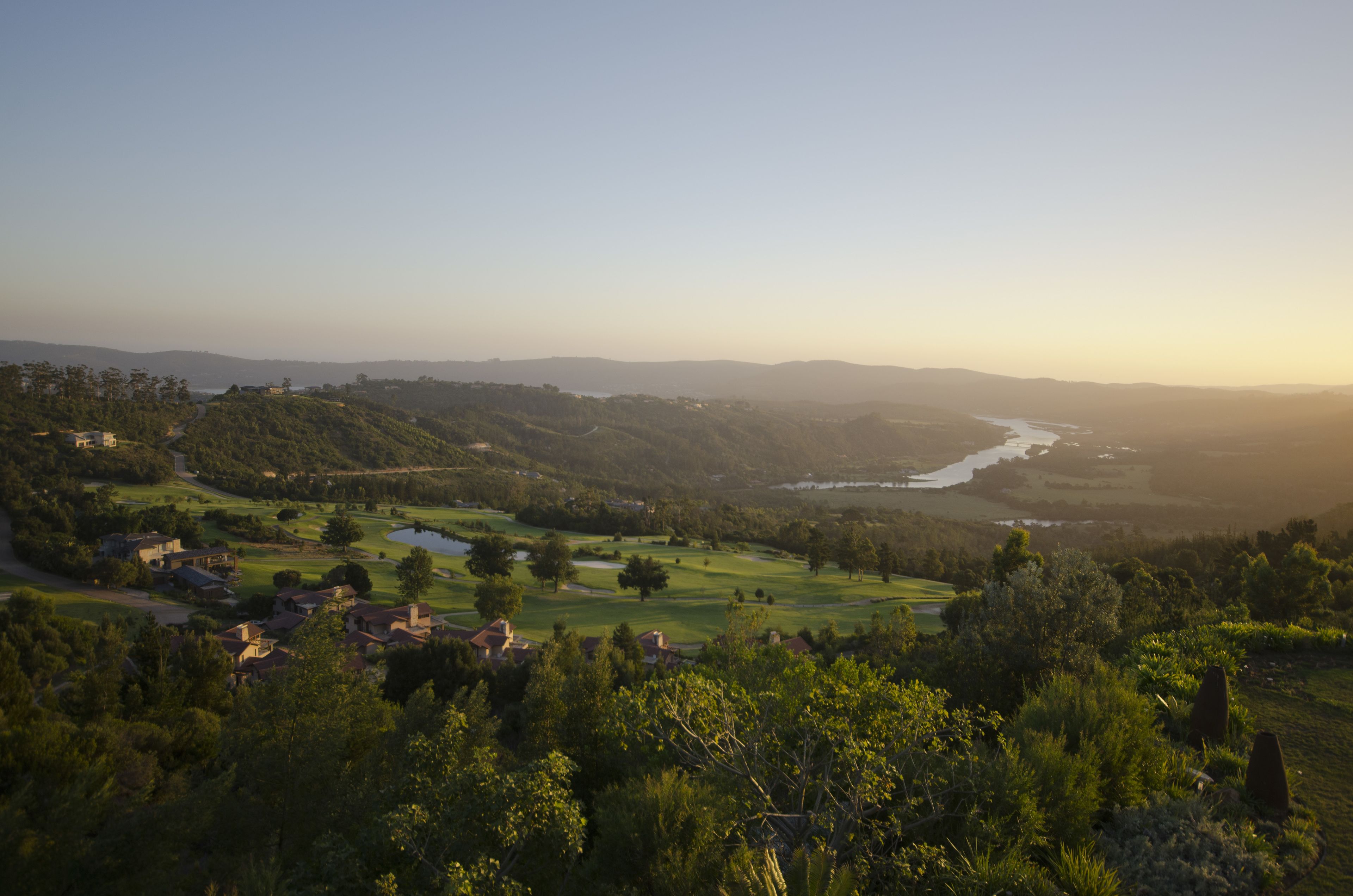 A panoramic view of a golf course with rolling hills, a river, and scattered buildings.