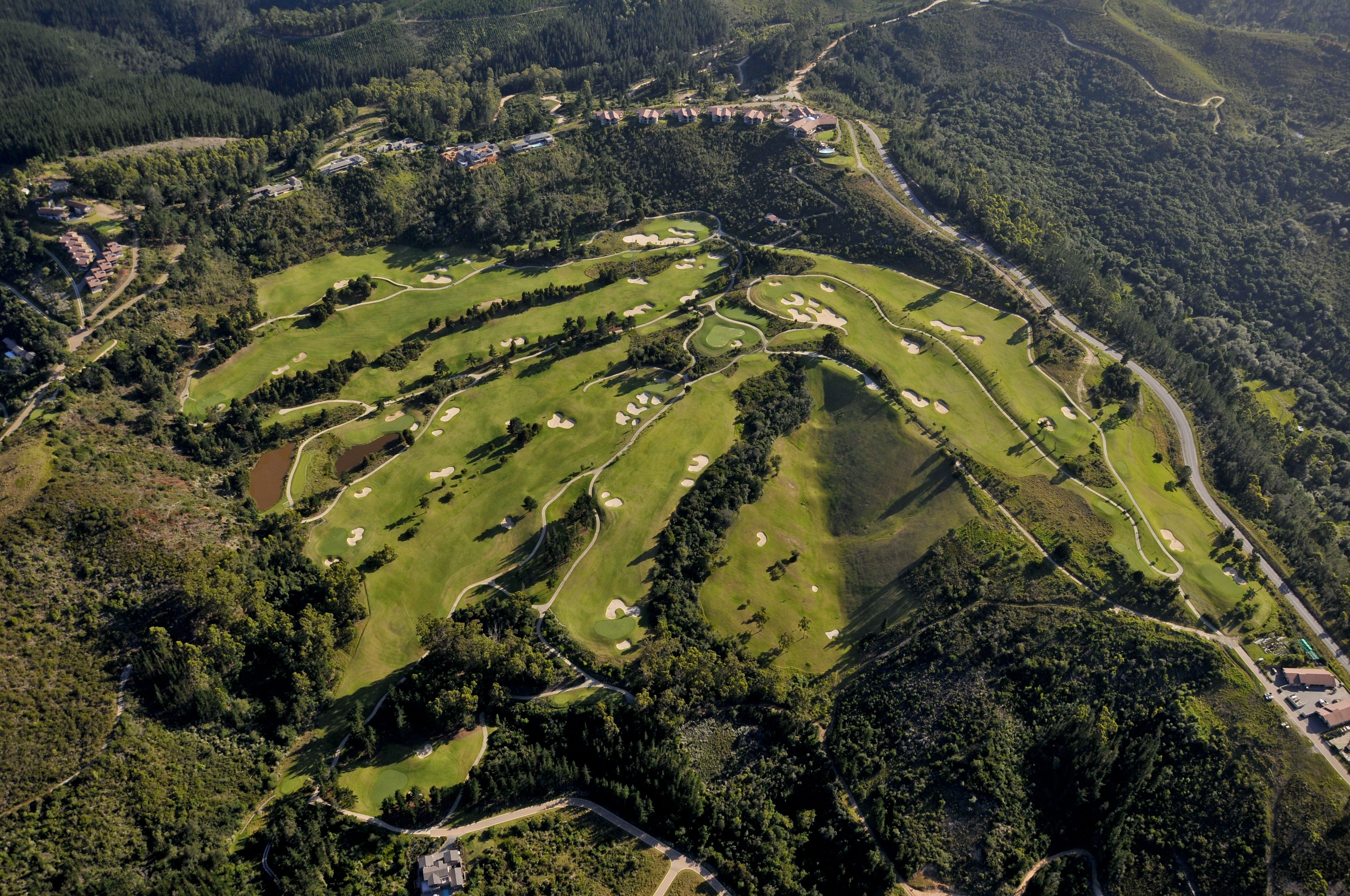 Vue aérienne d’un terrain de golf comportant plusieurs trous, entouré d’une forêt dense et de bâtiments résidentiels.