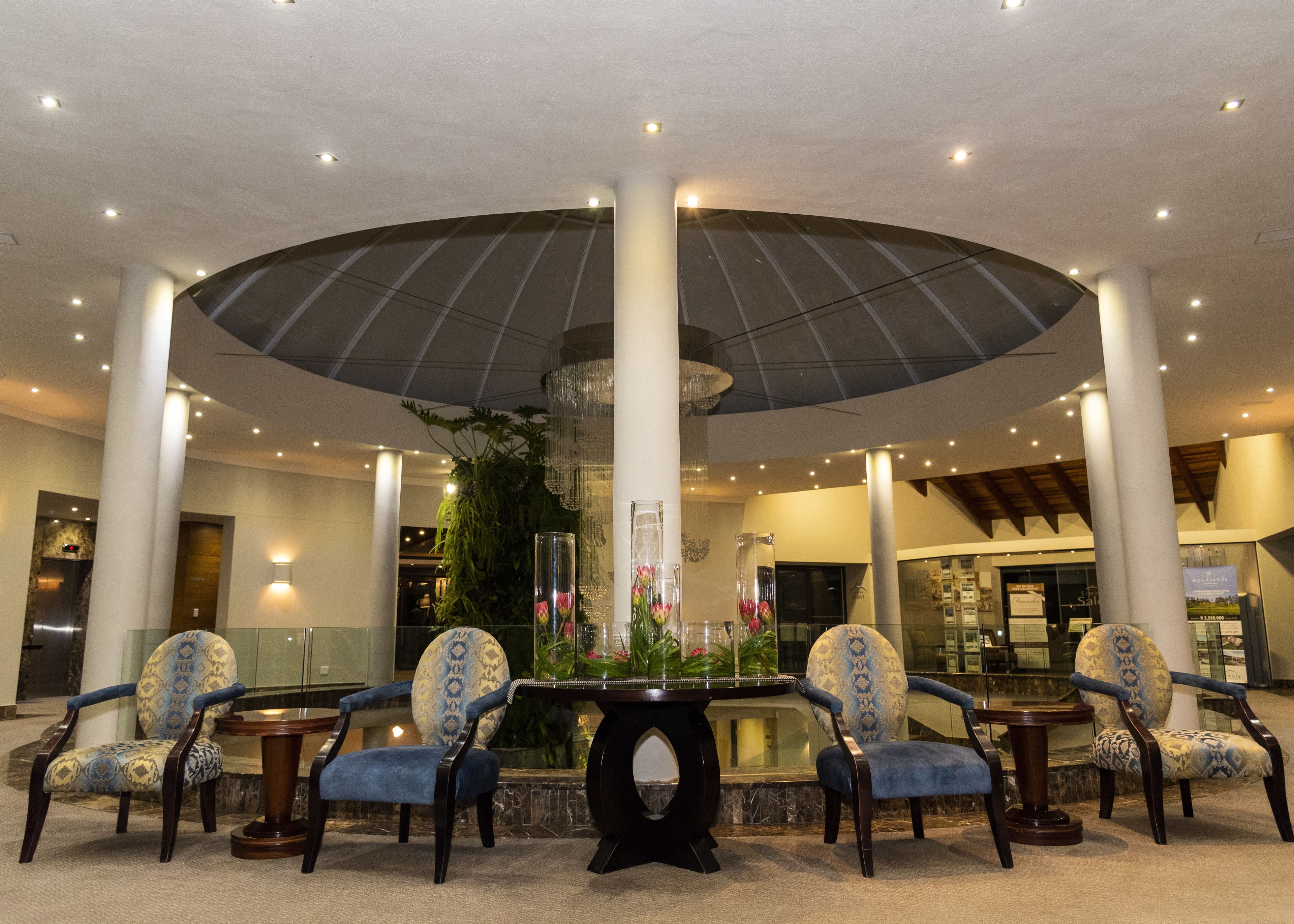 A lobby with a central round table surrounded by four patterned chairs, a large umbrella-like structure overhead, and a decorative plant.