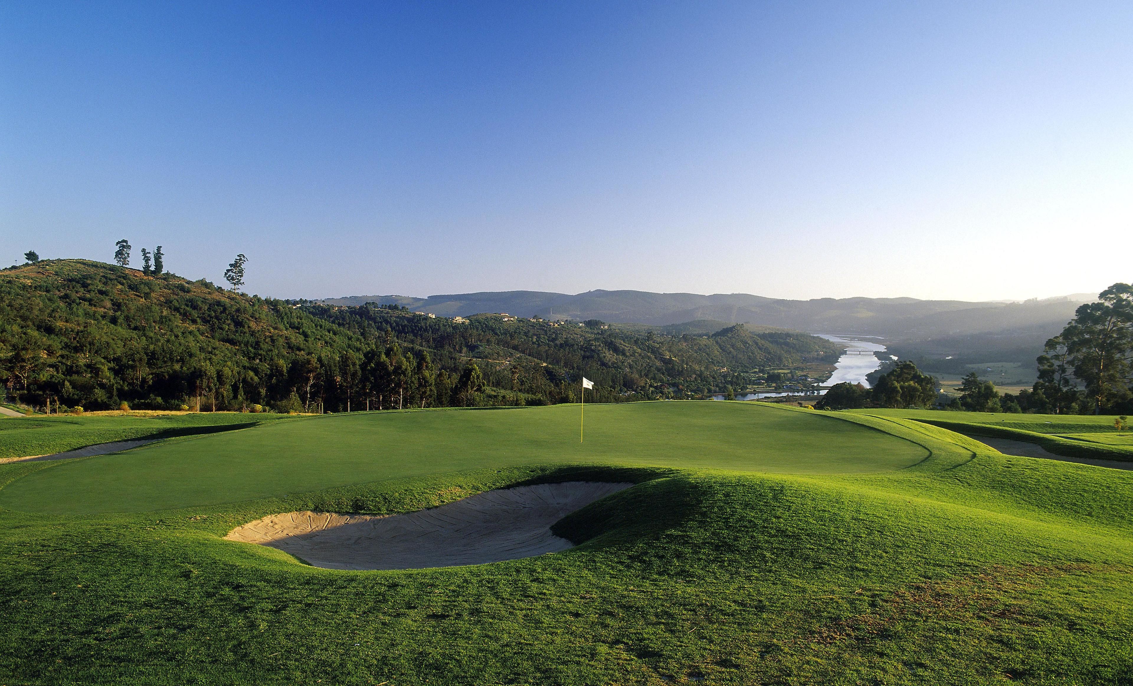 A golf course with a flag, sand bunkers, and a river in the background.