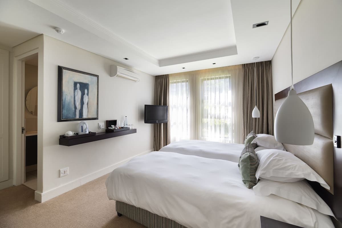 A hotel room with two beds, a flat-screen TV, a painting, a minibar, and a large window with curtains.