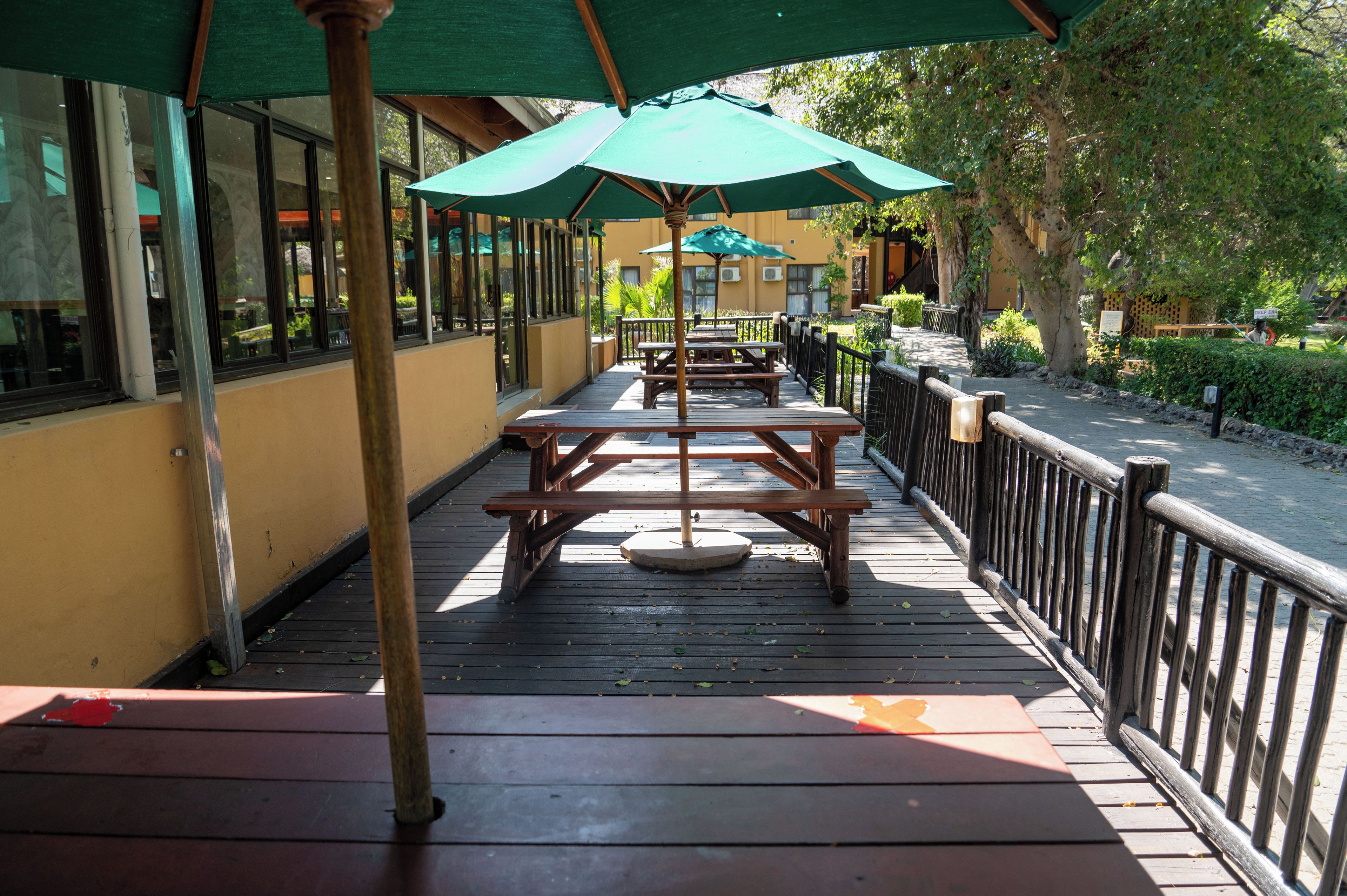 An outdoor patio with wooden tables and green umbrellas, surrounded by a wooden railing and a yellow building in the background.