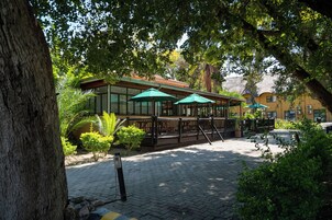 Un espacio al aire libre con mesas y sombrillas, rodeado de árboles y un edificio de fachada amarilla.