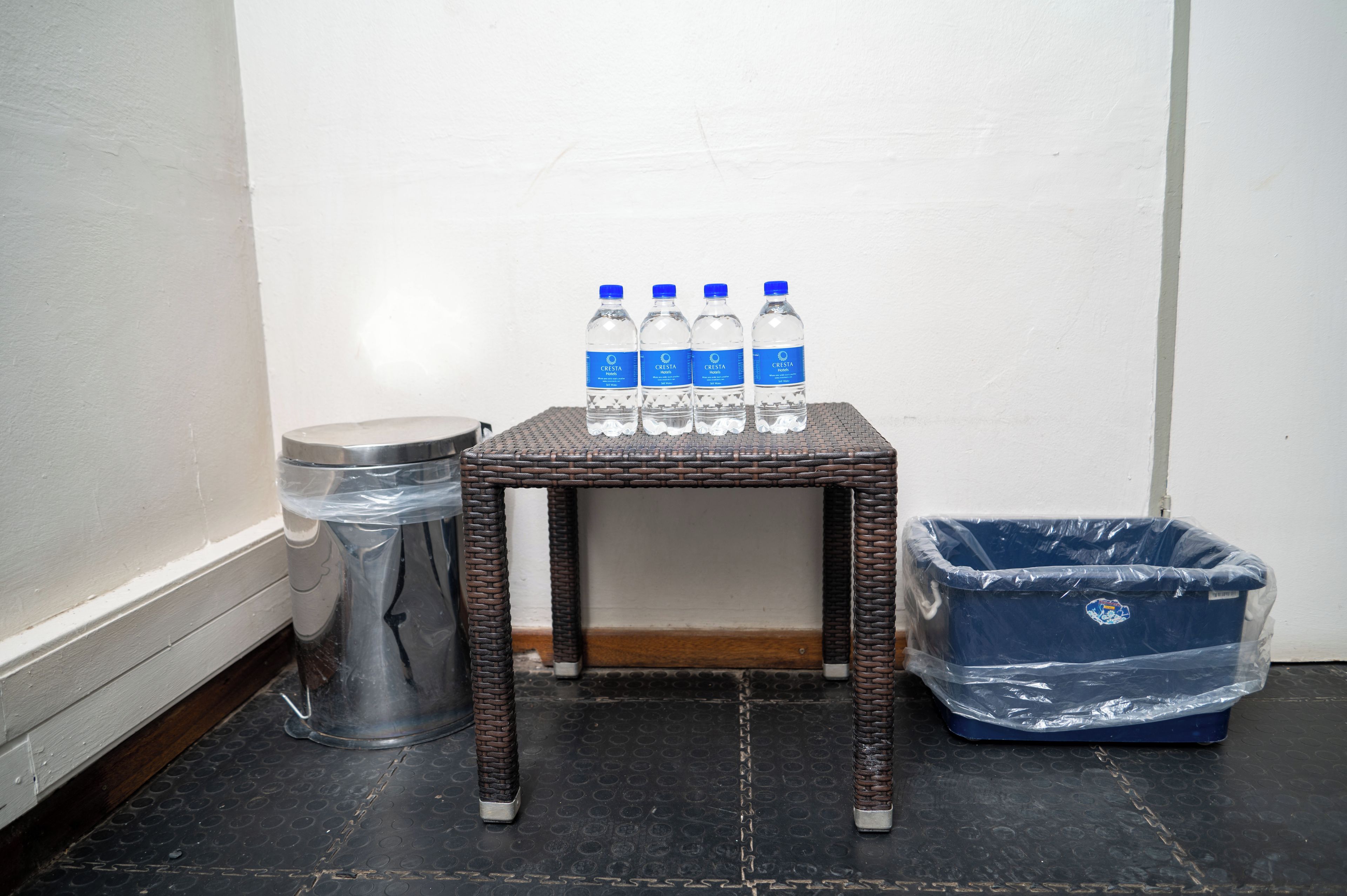 A small wicker table with three water bottles on top, a trash bin, and a covered waste bin.