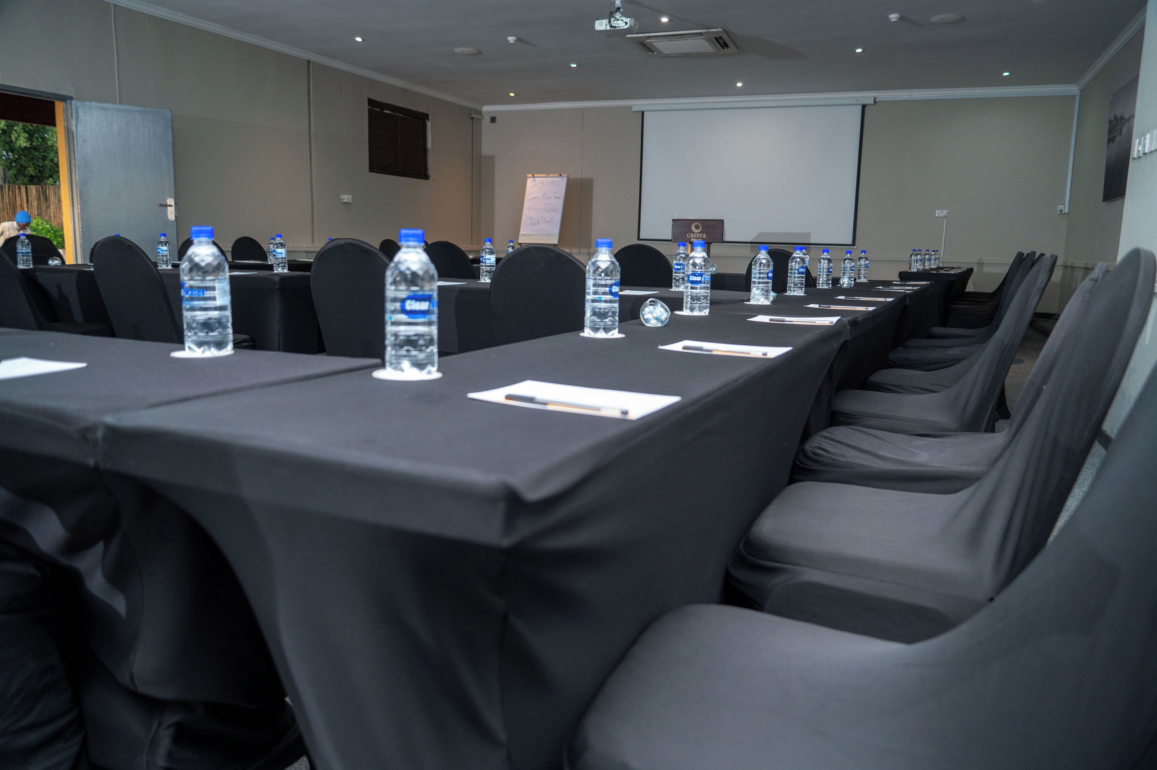 Un salón de conferencias con filas de mesas, sillas, botellas de agua y una pantalla de proyector.