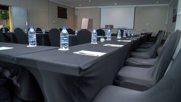 Un salón de conferencias con filas de mesas, sillas, botellas de agua y una pantalla de proyector.
