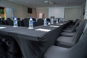 Un salón de conferencias con filas de mesas, sillas, botellas de agua y una pantalla de proyector.